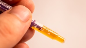 A close-up view of a person's fingers lightly holding a syringe filled with a yellow liquid. The syringe displays measurements in white against a purple and clear background.