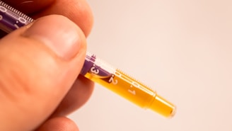 A close-up view of a person's fingers lightly holding a syringe filled with a yellow liquid. The syringe displays measurements in white against a purple and clear background.