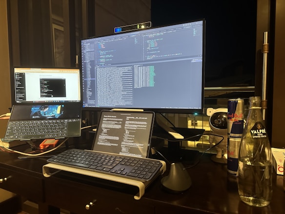 A desk setup for programming or coding, featuring multiple screens displaying code and text editors. A keyboard and a trackpad are positioned on the desk. There are also various items including a can of Red Bull, a bottle of Valpr&eacute; spring water, and a webcam placed next to the monitor.