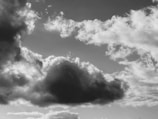 A black and white photograph of a sky filled with various clouds. A large, dark cloud is prominently centered with lighter, fluffy clouds surrounding it.