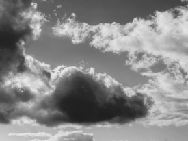 A black and white photograph of a sky filled with various clouds. A large, dark cloud is prominently centered with lighter, fluffy clouds surrounding it.