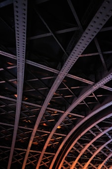 A detailed view of an iron structure with intersecting beams forming a lattice pattern. The beams are riveted and the entire structure is illuminated with a warm, orange light creating a dramatic contrast against the dark background.
