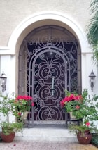 A decorative wrought iron security gate with intricate scrollwork stands in front of a wooden entry door. The archway above the gate complements the elegant design. Two wall-mounted lanterns flank the entrance, providing a classic touch. Bright red flowering plants in pots add a vibrant contrast to the neutral tones of the walls.