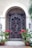 A decorative wrought iron security gate with intricate scrollwork stands in front of a wooden entry door. The archway above the gate complements the elegant design. Two wall-mounted lanterns flank the entrance, providing a classic touch. Bright red flowering plants in pots add a vibrant contrast to the neutral tones of the walls.