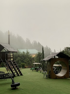 A misty outdoor playground area is surrounded by lush green trees. The structures include wooden playhouses and swings set against a backdrop of fog-covered trees. There is a roofed seating area with intricate designs and a circular tunnel-like play structure.
