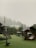 A misty outdoor playground area is surrounded by lush green trees. The structures include wooden playhouses and swings set against a backdrop of fog-covered trees. There is a roofed seating area with intricate designs and a circular tunnel-like play structure.