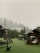 A misty outdoor playground area is surrounded by lush green trees. The structures include wooden playhouses and swings set against a backdrop of fog-covered trees. There is a roofed seating area with intricate designs and a circular tunnel-like play structure.