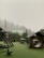 A misty outdoor playground area is surrounded by lush green trees. The structures include wooden playhouses and swings set against a backdrop of fog-covered trees. There is a roofed seating area with intricate designs and a circular tunnel-like play structure.