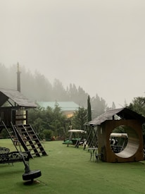 A misty outdoor playground area is surrounded by lush green trees. The structures include wooden playhouses and swings set against a backdrop of fog-covered trees. There is a roofed seating area with intricate designs and a circular tunnel-like play structure.