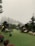 A foggy outdoor scene features a playground with wooden structures, including slides and climbing frames. The area is surrounded by lush trees, and the ground is covered with artificial grass. Several decorative sheep statues are placed near the play area, and there are various benches and potted plants around.