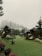 A foggy outdoor scene features a playground with wooden structures, including slides and climbing frames. The area is surrounded by lush trees, and the ground is covered with artificial grass. Several decorative sheep statues are placed near the play area, and there are various benches and potted plants around.