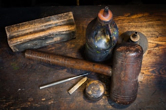 A collection of antique woodworking tools is arranged on a rustic wooden surface. The tools include a large wooden mallet, a smaller metal tool resembling a chisel, and two vintage oil cans with a worn, weathered appearance. The surface of the wood is aged, adding to the vintage atmosphere of the scene.