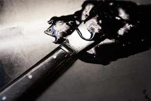 A paint spatula with a dark wooden handle is resting on a surface covered with a shiny black liquid. The light reflects off the smooth, glossy liquid and the metallic part of the spatula.