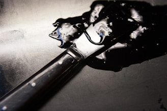 A paint spatula with a dark wooden handle is resting on a surface covered with a shiny black liquid. The light reflects off the smooth, glossy liquid and the metallic part of the spatula.