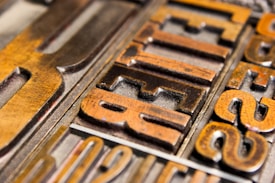 Close-up of vintage metal letterpress blocks arranged with visible woodgrain texture. The letters appear worn, indicating age and use, and are in various shades of brown and gold.