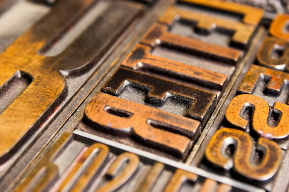 Close-up of vintage metal letterpress blocks arranged with visible woodgrain texture. The letters appear worn, indicating age and use, and are in various shades of brown and gold.