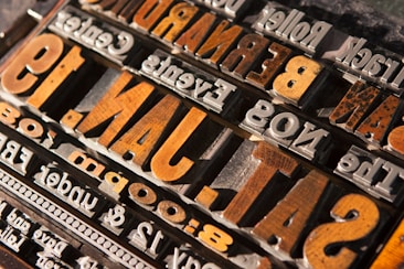 Wooden and metal printing blocks are arranged to form backward and upside-down text, featuring large letters and numbers with a vintage style appearance. The textures of the blocks give a tactile sense, with some blocks showing signs of wear and age.