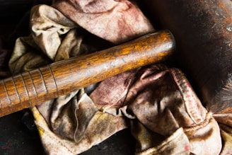A weathered wooden bat rests on a bundle of old, stained cloth with a dark leather object nearby. The textures are rustic and aged.