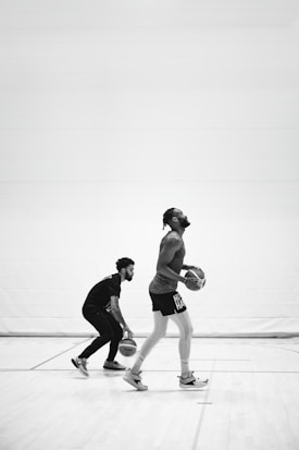 Two men are on a basketball court; one dribbles a ball while another prepares to shoot. The scene is captured in black and white, emphasizing the focus and motion.