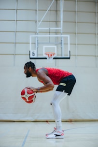 A basketball player in a red jersey and black shorts is practicing dribbling on an indoor court. The player is focused, holding two basketballs, with a basketball hoop and court lines visible in the background.