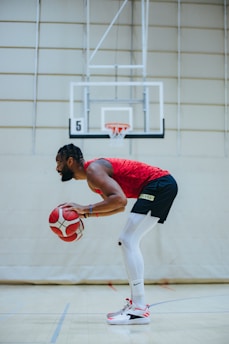 A basketball player in a red jersey and black shorts is practicing dribbling on an indoor court. The player is focused, holding two basketballs, with a basketball hoop and court lines visible in the background.