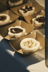 Delicately arranged desserts are placed in square, brown paper trays. Each dessert is round with a white, sponge-like outer layer and different toppings on top, such as chocolate and nuts. They are displayed on a white surface, partially lit by warm light.