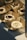 Delicately arranged desserts are placed in square, brown paper trays. Each dessert is round with a white, sponge-like outer layer and different toppings on top, such as chocolate and nuts. They are displayed on a white surface, partially lit by warm light.