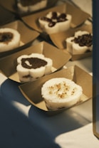 Delicately arranged desserts are placed in square, brown paper trays. Each dessert is round with a white, sponge-like outer layer and different toppings on top, such as chocolate and nuts. They are displayed on a white surface, partially lit by warm light.