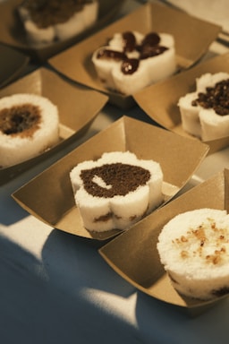 A series of desserts arranged in individual brown paper trays. Each dessert is a round, layered piece with both white and dark brown sections, possibly made of steamed rice flour and brown sugar. Some are topped with red bean paste and others with small granular toppings.