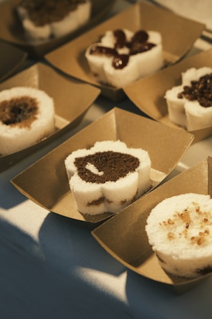 A series of desserts arranged in individual brown paper trays. Each dessert is a round, layered piece with both white and dark brown sections, possibly made of steamed rice flour and brown sugar. Some are topped with red bean paste and others with small granular toppings.