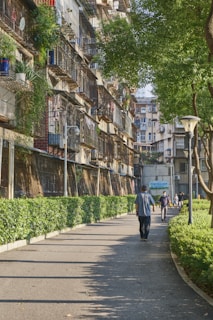 A residential area with several multi-story apartment buildings featuring balconies with metal grilles. Greenery is abundant, with trees and well-maintained hedges lining a paved pathway. People are casually strolling down the walkway, creating a serene and livable atmosphere.