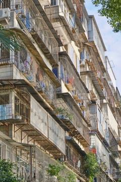 A tall apartment building with multiple floors featuring balconies protected by metal bars. The balconies have various items such as potted plants, clothes, and satellite dishes. The structure is predominantly made of concrete, and some of the apartments have air conditioning units installed. Vegetation can be seen around and below the building.