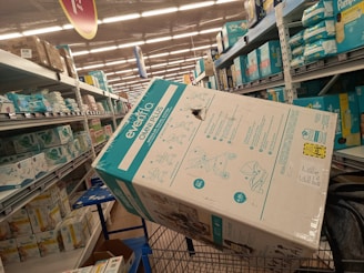 A shopping aisle filled with shelves stacked with boxes of diapers. A large box labeled 'Evenflo Omni Plus' is prominently placed in a shopping cart. Other diaper brands such as Pampers are visible on the shelves. The setting appears to be a supermarket or a retail store with fluorescent lighting.