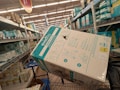 A shopping aisle filled with shelves stacked with boxes of diapers. A large box labeled 'Evenflo Omni Plus' is prominently placed in a shopping cart. Other diaper brands such as Pampers are visible on the shelves. The setting appears to be a supermarket or a retail store with fluorescent lighting.