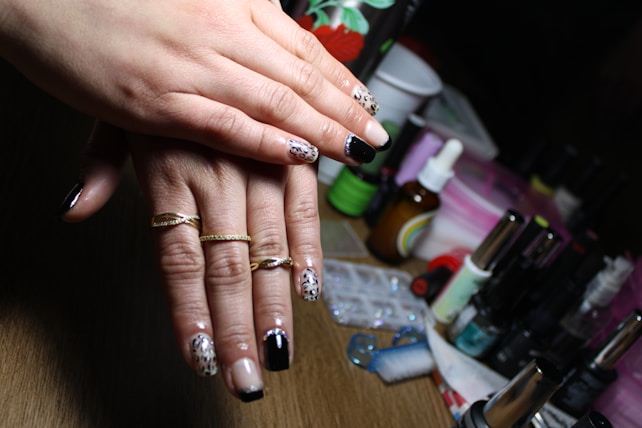 A pair of hands with polished nails featuring intricate designs, including leopard print and glossy black, adorned with gold rings. In the background, various manicure tools and nail polish bottles are arranged on a table, along with a dropper bottle and other beauty products.