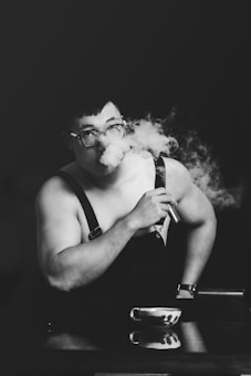 A person with short hair and glasses is exhaling vapor from an electronic cigarette. They are wearing dark overalls and a watch, leaning on a surface with a small cup in front. The scene is captured in black and white, creating a smoky, artistic atmosphere.