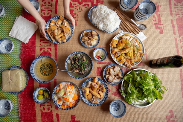 A variety of traditional dishes are laid out on a woven mat, including bowls of soup, plates of cooked meats, vegetables, rice noodles, herbs, and dipping sauces. A pair of hands is placing a plate of food on the mat. There are also cups, chopsticks, a bottle, and a block of tofu or cake in the scene.