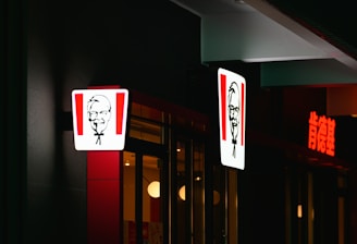 A brightly lit restaurant sign featuring a man's face with glasses, bowtie, and goatee is visible. The scene includes illuminated signs, part of a glass storefront with reflections, minimal interior lighting, and a dark surrounding exterior.