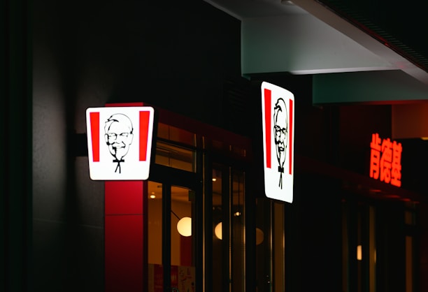 A brightly lit restaurant sign featuring a man's face with glasses, bowtie, and goatee is visible. The scene includes illuminated signs, part of a glass storefront with reflections, minimal interior lighting, and a dark surrounding exterior.