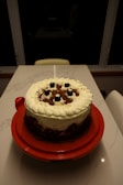 A round cake with a layer of whipped cream frosting and chocolate shavings, topped with small dark cherries. The cake is placed on a red cake stand on a marble countertop, with a dimly lit background featuring a window and a chair.