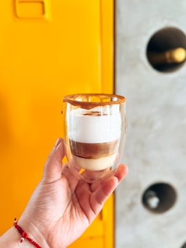 A hand with a red and white beaded bracelet holds a double-walled glass containing a layered coffee beverage. The layers include milk, coffee, and foam, topped with a brown sugar or cinnamon rim. The background features a bright yellow cabinet and a gray wall with circular cutouts.