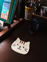 A brown wooden surface holds a cat-shaped mouse pad with a cartoon face featuring whiskers and stripes. Nearby, a desk organizer contains pencils, art supplies, and a cup with yellow drips. A black Sony speaker is visible, and a book with colorful cartoon inserts rests on a wooden stand.