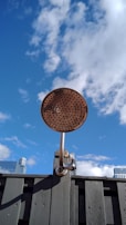 A metallic outdoor shower head is mounted on a wooden fence against a backdrop of a clear blue sky with scattered clouds. The top of some modern buildings can be seen in the distance.