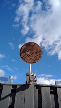 A metallic outdoor shower head is mounted on a wooden fence against a backdrop of a clear blue sky with scattered clouds. The top of some modern buildings can be seen in the distance.