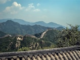 A scenic view of the Great Wall of China meandering over the lush green hills with a background of hazy blue mountains under a partly cloudy sky. The foreground includes traditional roof tiles, possibly from a nearby structure.