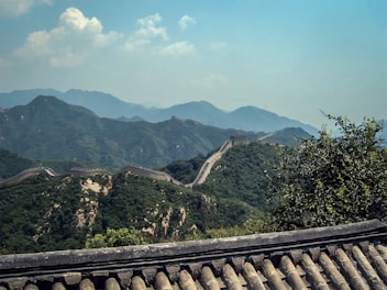 A scenic view of the Great Wall of China meandering over the lush green hills with a background of hazy blue mountains under a partly cloudy sky. The foreground includes traditional roof tiles, possibly from a nearby structure.