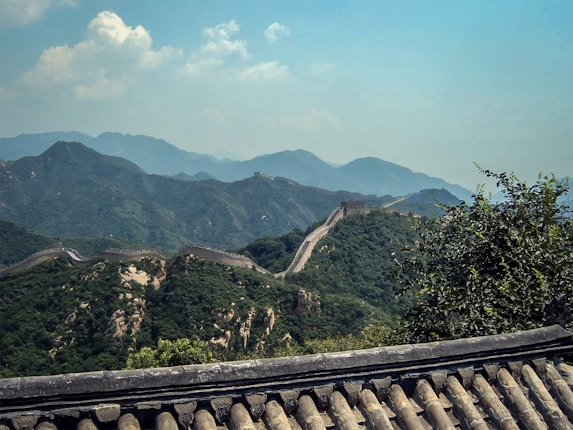 A scenic view of the Great Wall of China meandering over the lush green hills with a background of hazy blue mountains under a partly cloudy sky. The foreground includes traditional roof tiles, possibly from a nearby structure.