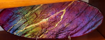 An oval-shaped glass object with a textured surface exhibits vibrant colors including deep purples, greens, and yellows. The surface has a glossy finish and features several wavy metallic gold lines running across it. It is positioned on a wooden display stand or shelf.