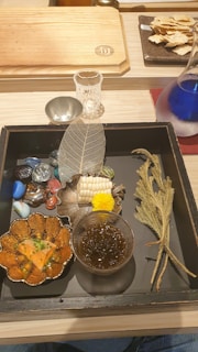 A beautifully arranged tray with a variety of food items including a dish with orange slices garnished with green herbs, a glass with a dark liquid, and decorative elements such as a translucent leaf and colorful stones. To the side, a small carafe contains a blue liquid.