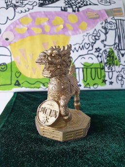 A metallic gold statue of a mythical creature stands on a hexagonal base. The creature holds a circular coin with inscriptions in its raised paw. Behind the statue is a colorful drawing with abstract elements including a large, pink and yellow fish-like figure, green grass, and various scribbles and shapes.